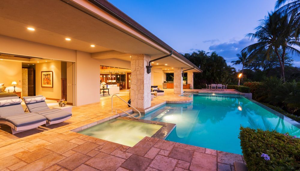 Poolside patio with pool, hot tub, and house. Blue pool water, lounge chairs, palm trees.