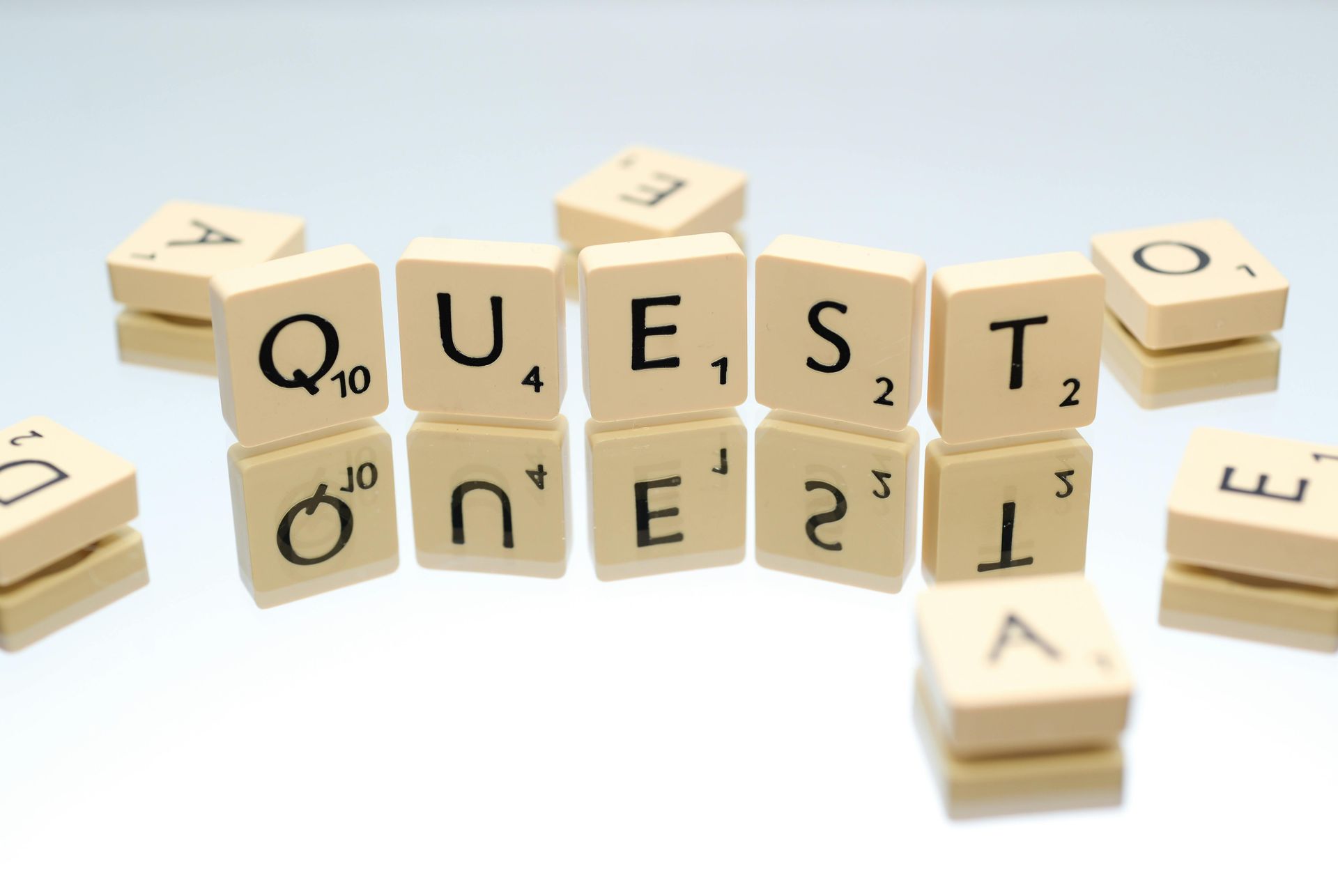 Scrabble tiles spelling QUEST with scattered tiles. White background.