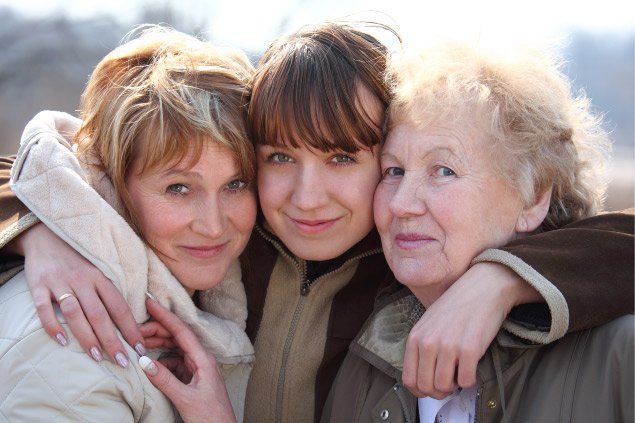 Three women of different generations smiling and embracing outdoors.