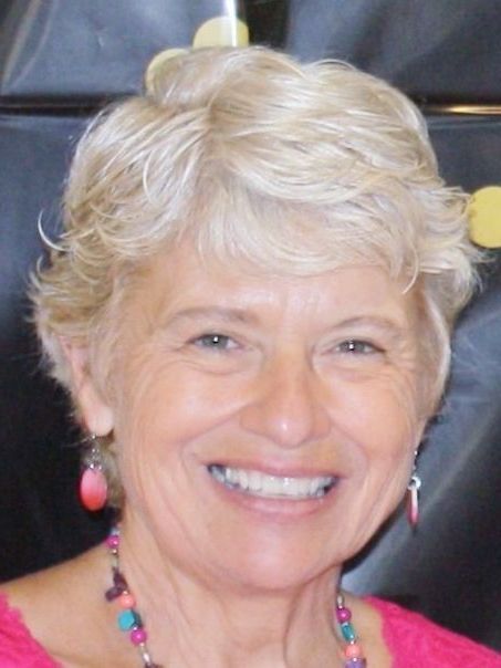 Smiling older woman with short, blonde hair, wearing pink earrings and a necklace, close-up.