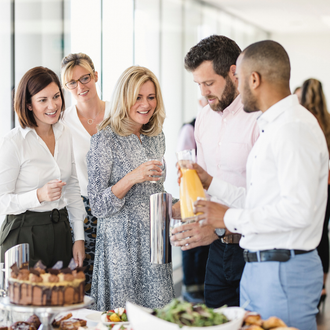 A diverse group of colleagues talking and laughing while enjoying drinks and food at an indoor office social gathering.
