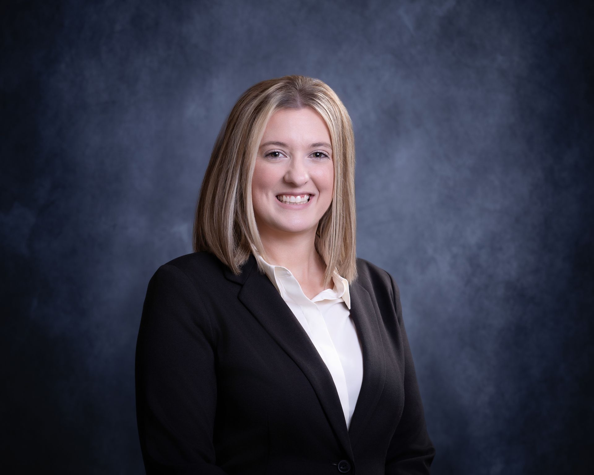 Professional headshot of a smiling person in a black blazer and white shirt against a dark studio backdrop