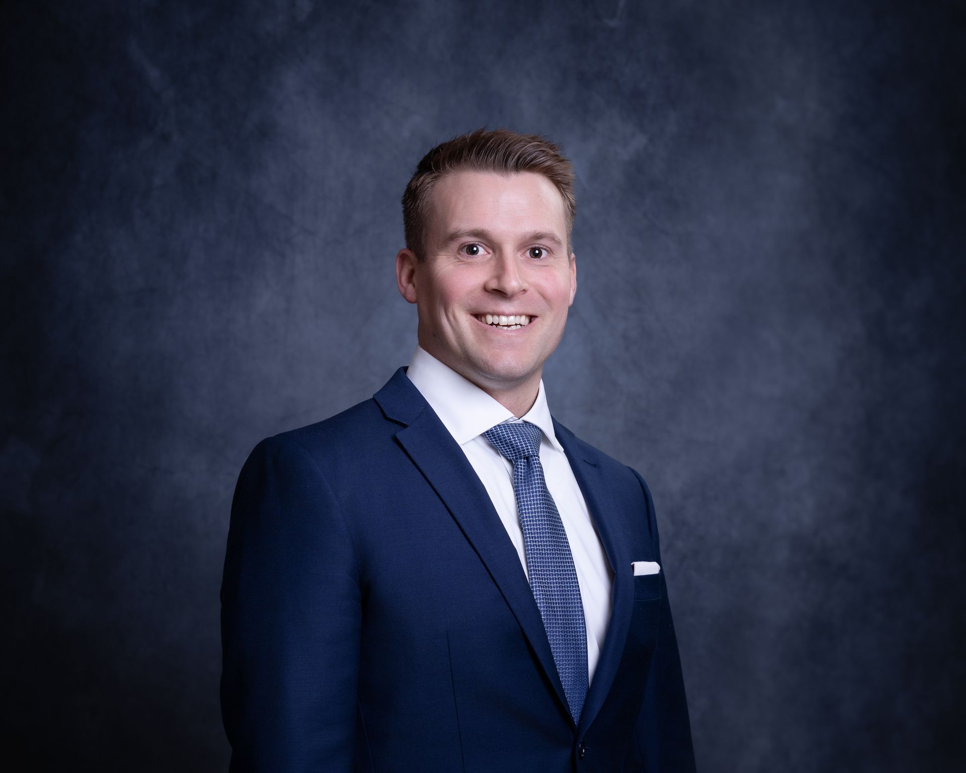 Studio portrait of a smiling man in a navy suit and tie against a dark gray backdrop