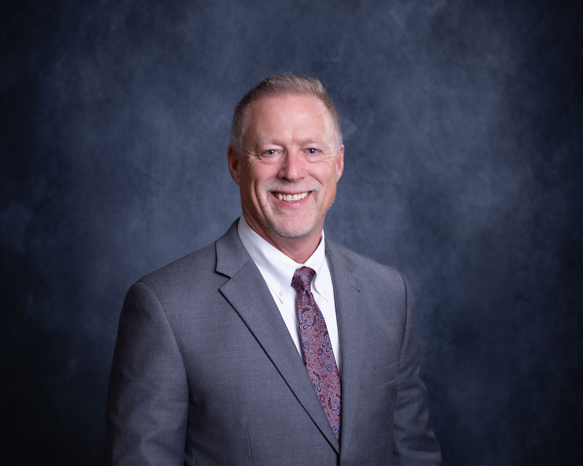 Smiling man in a gray suit with a patterned tie against a dark studio backdrop