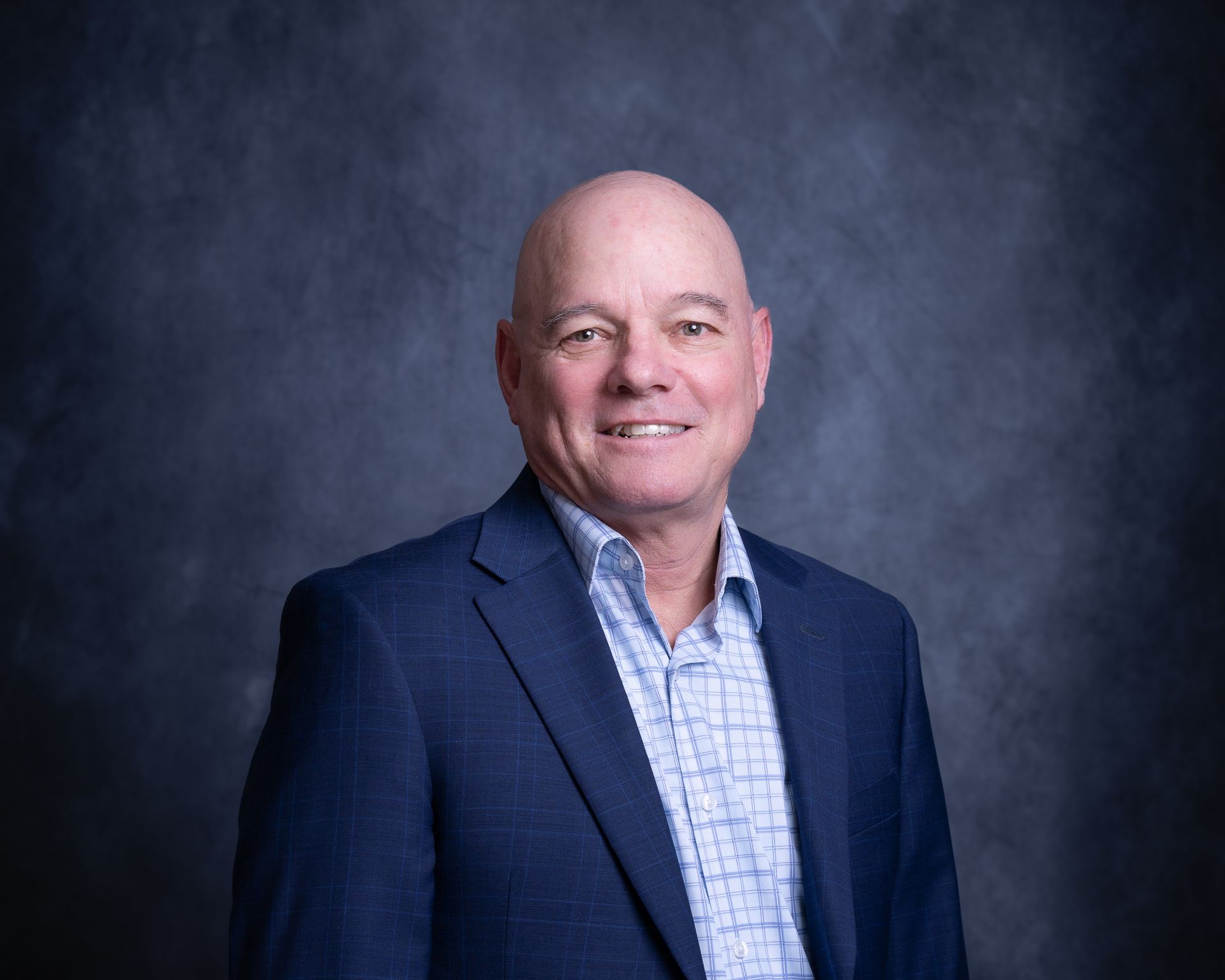 Portrait of a smiling bald man in a navy blazer and patterned shirt against a dark studio backdrop