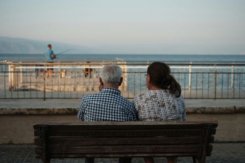 Two people sit on a bench facing the sea at a waterfront railing, with a distant cyclist and mountains beyond.