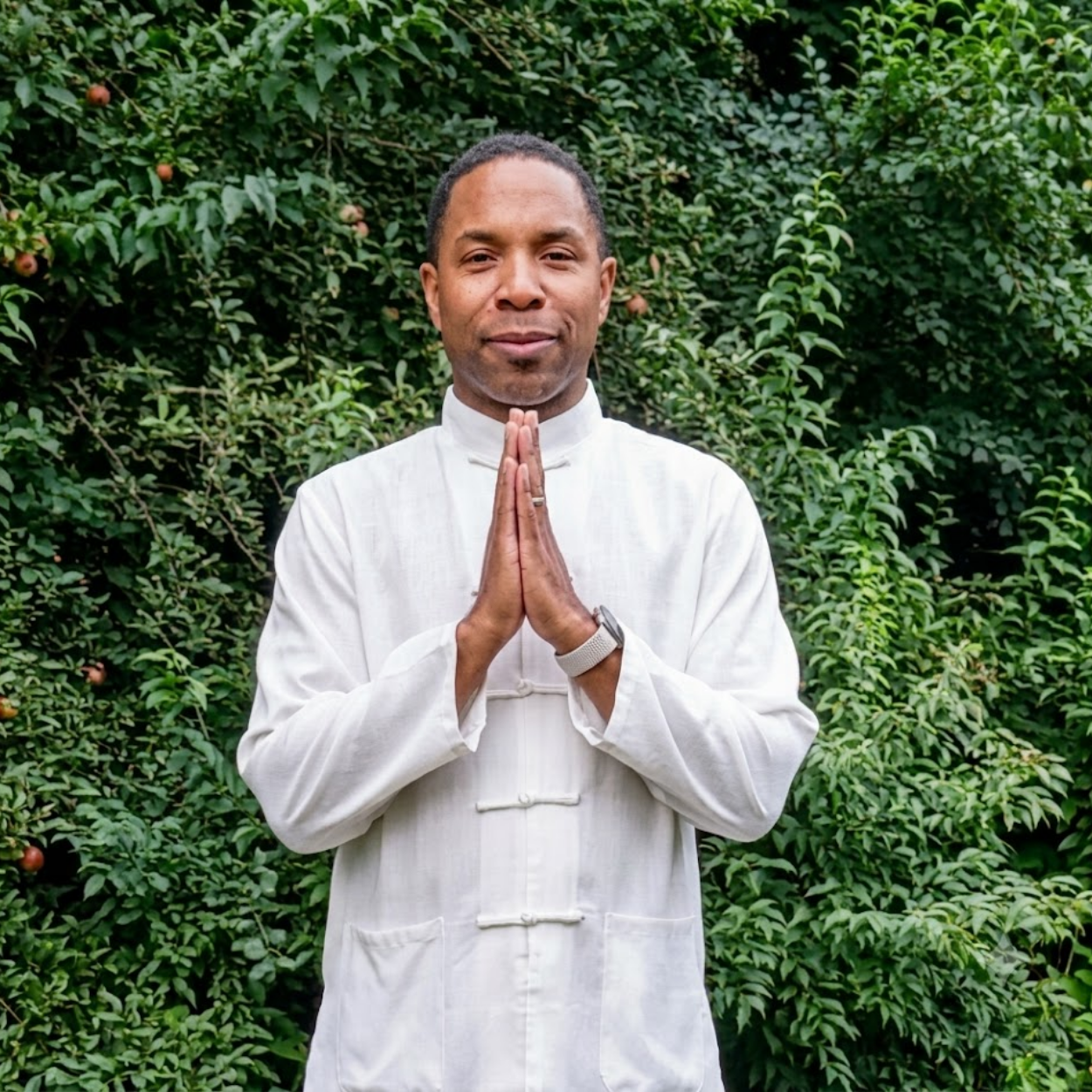 Man in white traditional clothing standing with hands together in front of green foliage
