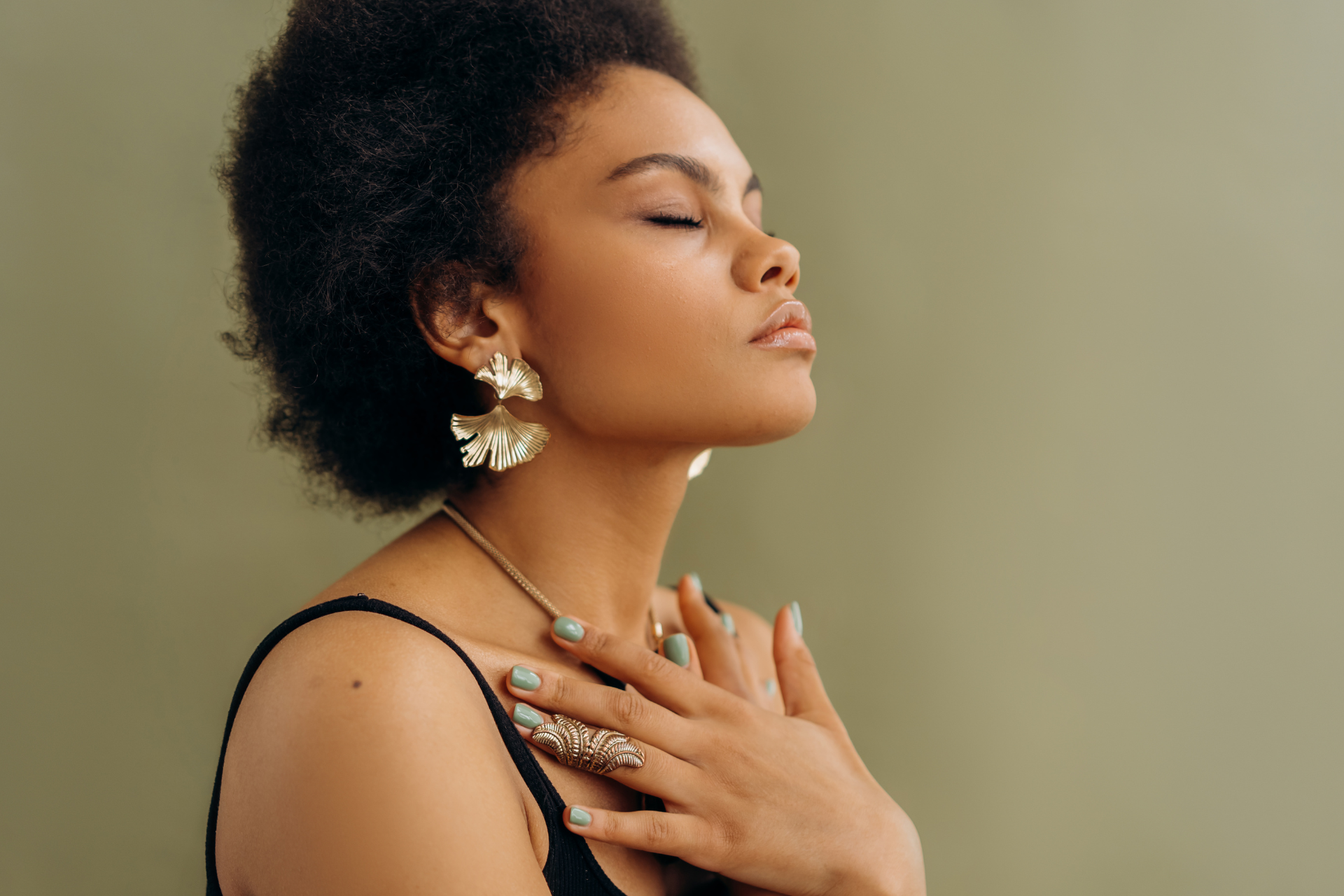 Woman in a black dress with gold jewelry, eyes closed, hands on chest against a beige background