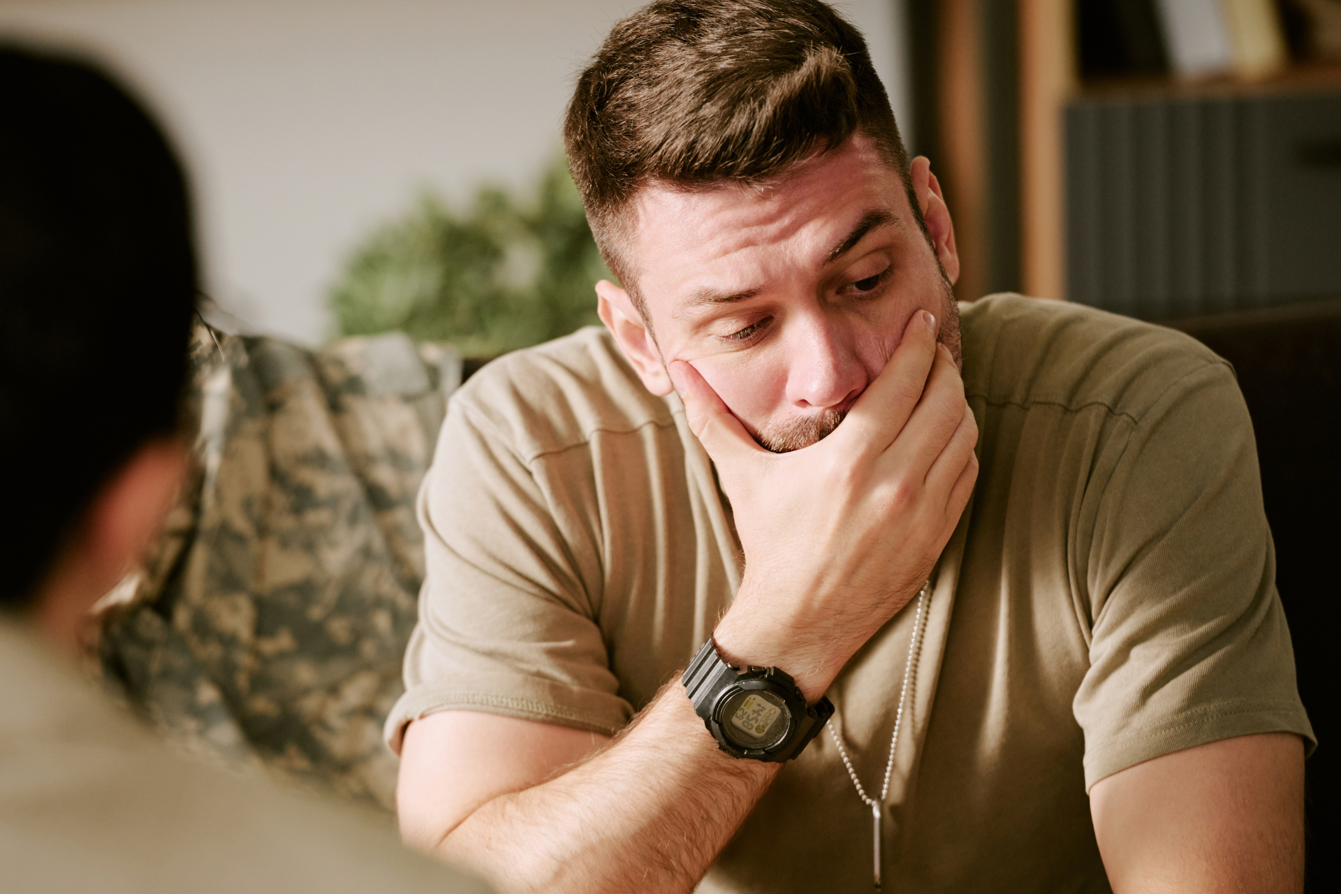 Man in beige shirt sitting with hand over mouth, looking down pensively in a room
