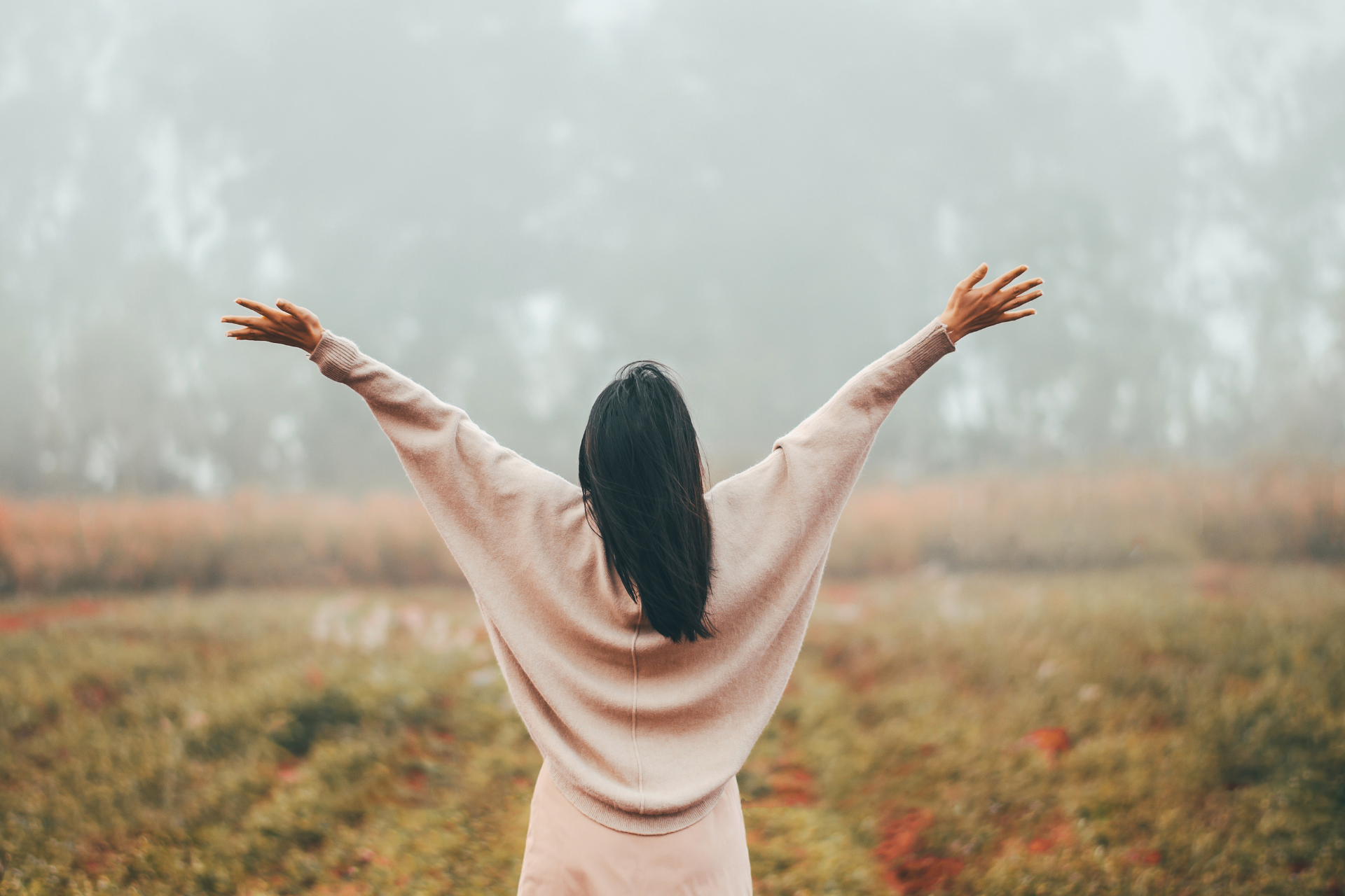 Person with raised arms standing in a misty field, facing away in a beige sweater