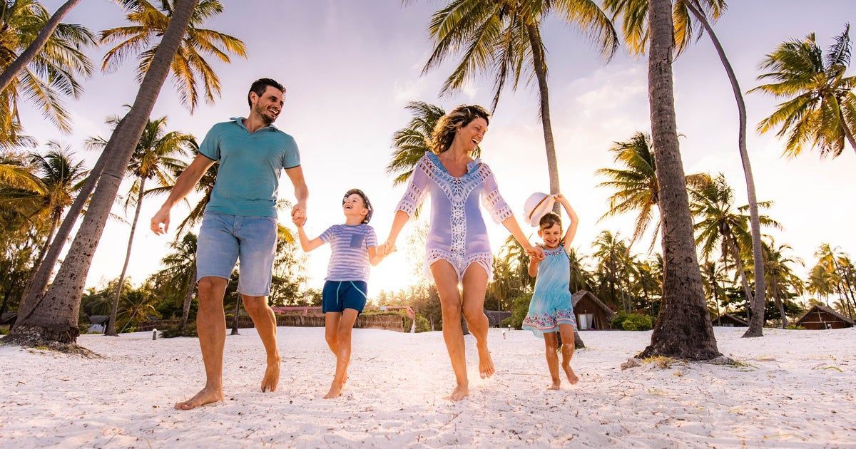 Family holding hands, walking on a sandy beach under palm trees, sun shining.