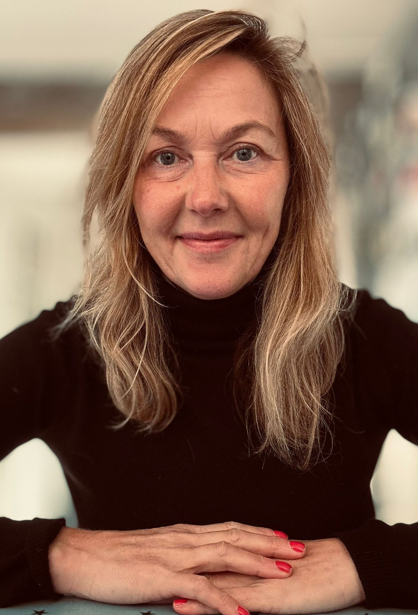 A woman in a black turtleneck is sitting at a table with her hands folded.