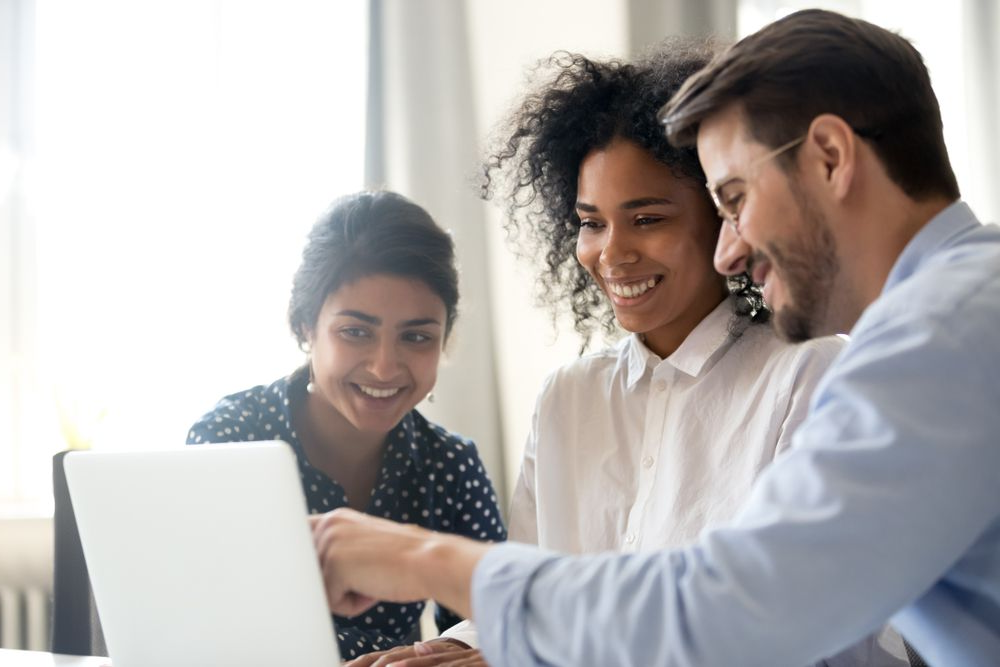 Three professionals smile while looking at a laptop screen, with one person pointing at the display.
