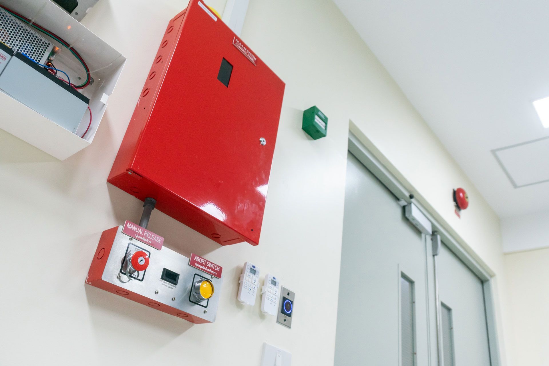 Red emergency cabinet and controls beside a gray door in a white hallway