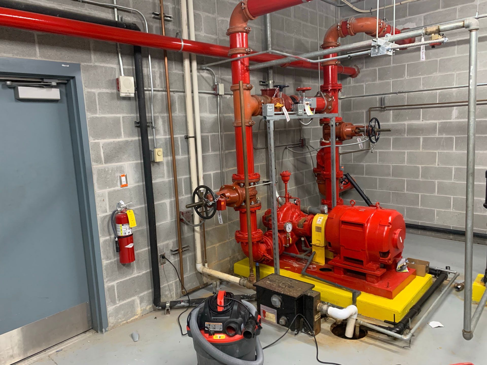 Red fire pump and piping in a concrete utility room beside a gray door.