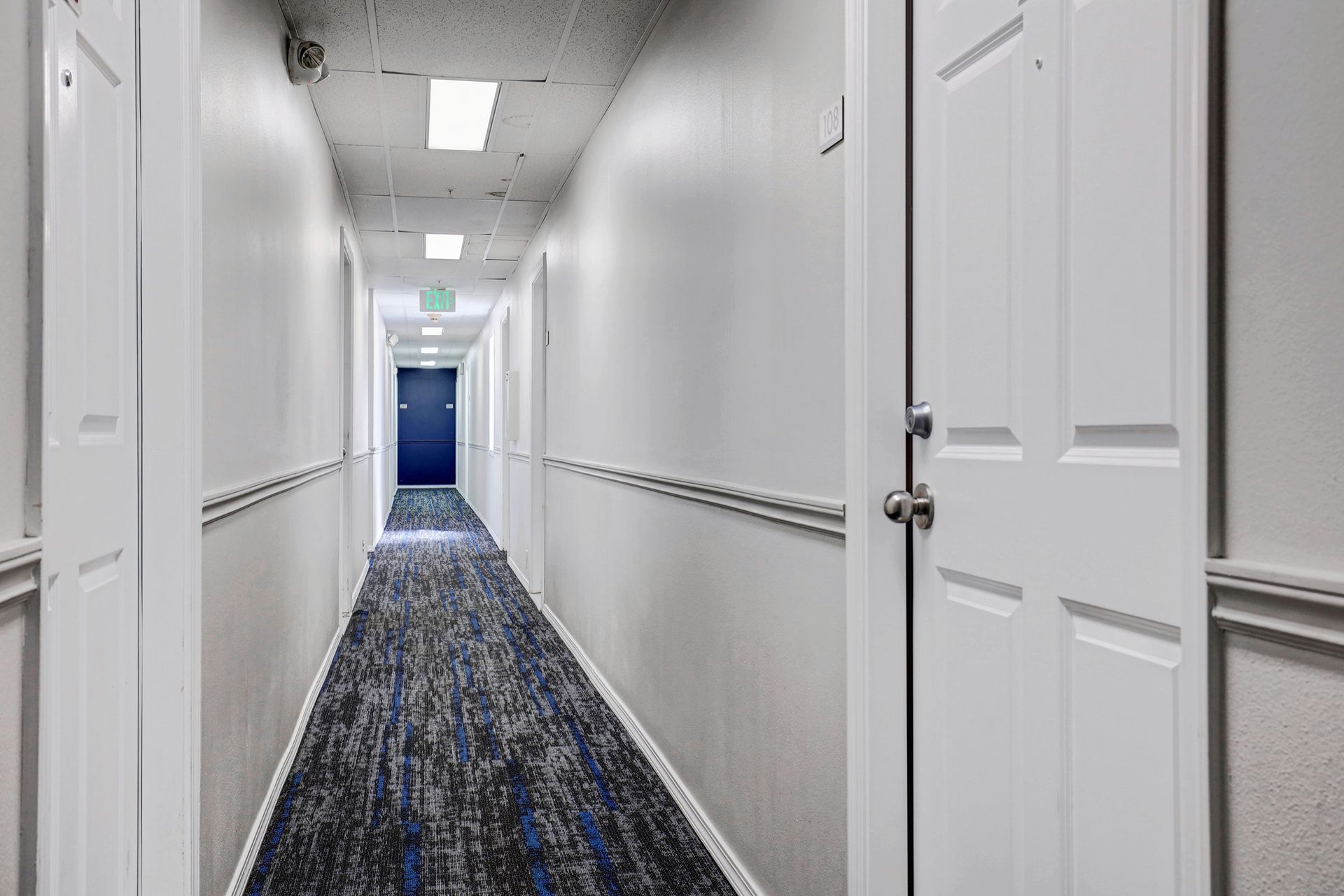 A long hallway with white walls and blue carpet and a blue door.