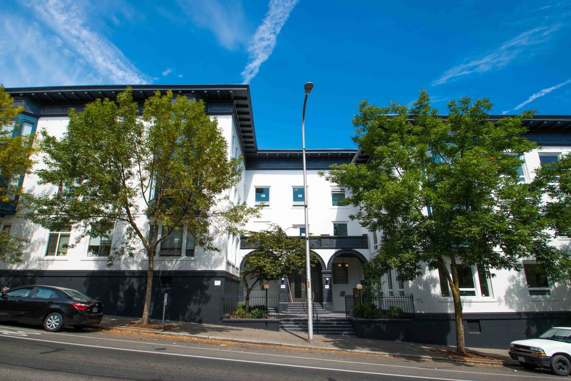 A large white building with trees in front of it and cars parked in front of it.