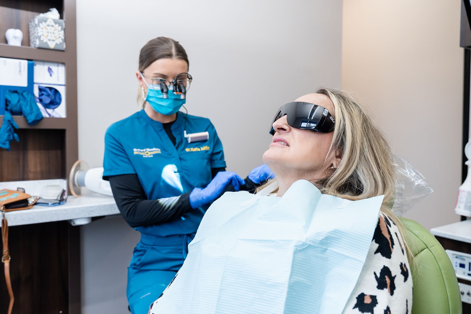 A dental professional in blue scrubs and a face mask examining a patient wearing protective eyewear in a clinical office.
