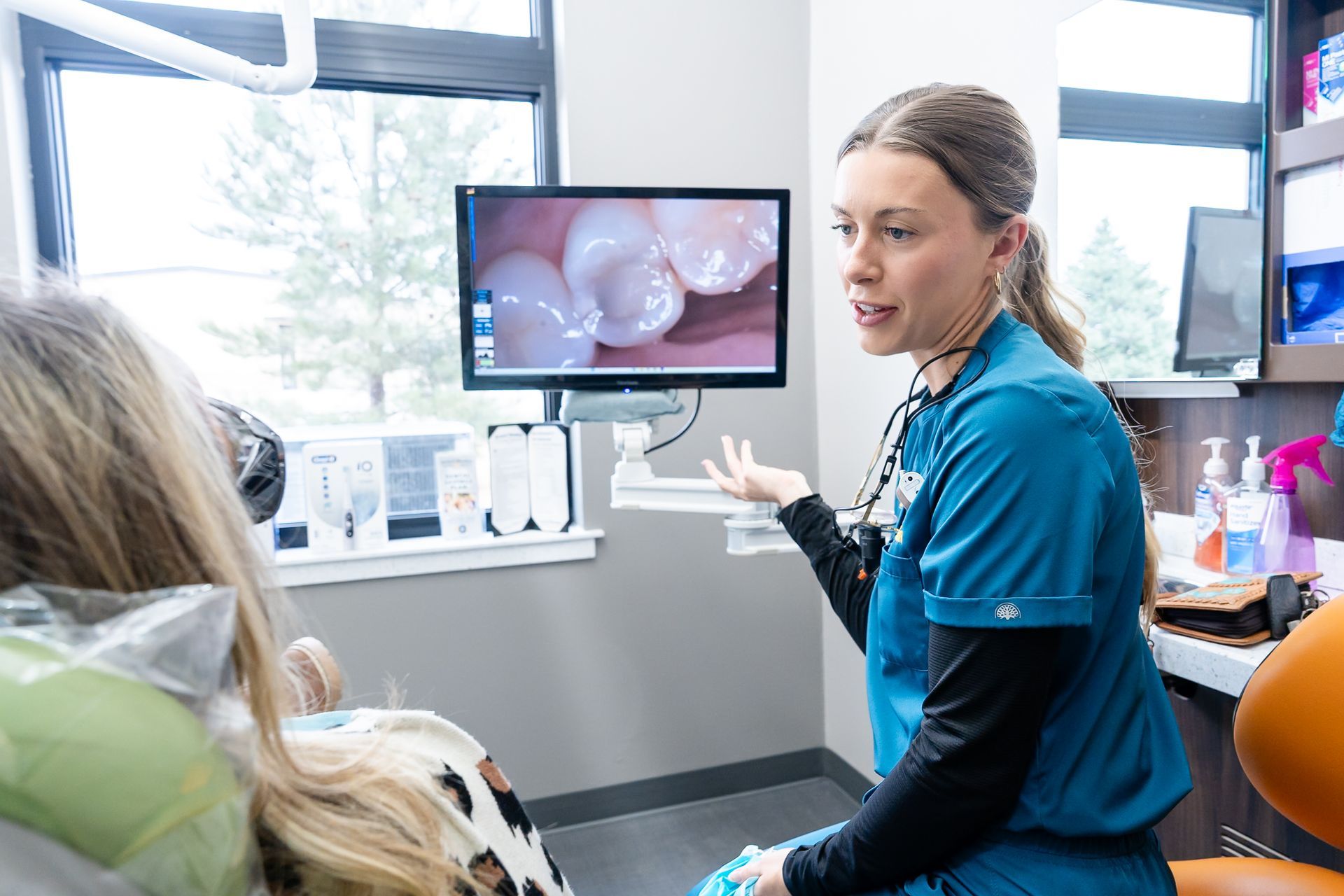 A dental professional in teal scrubs sits and talks with a patient reclined in a green dental chair.