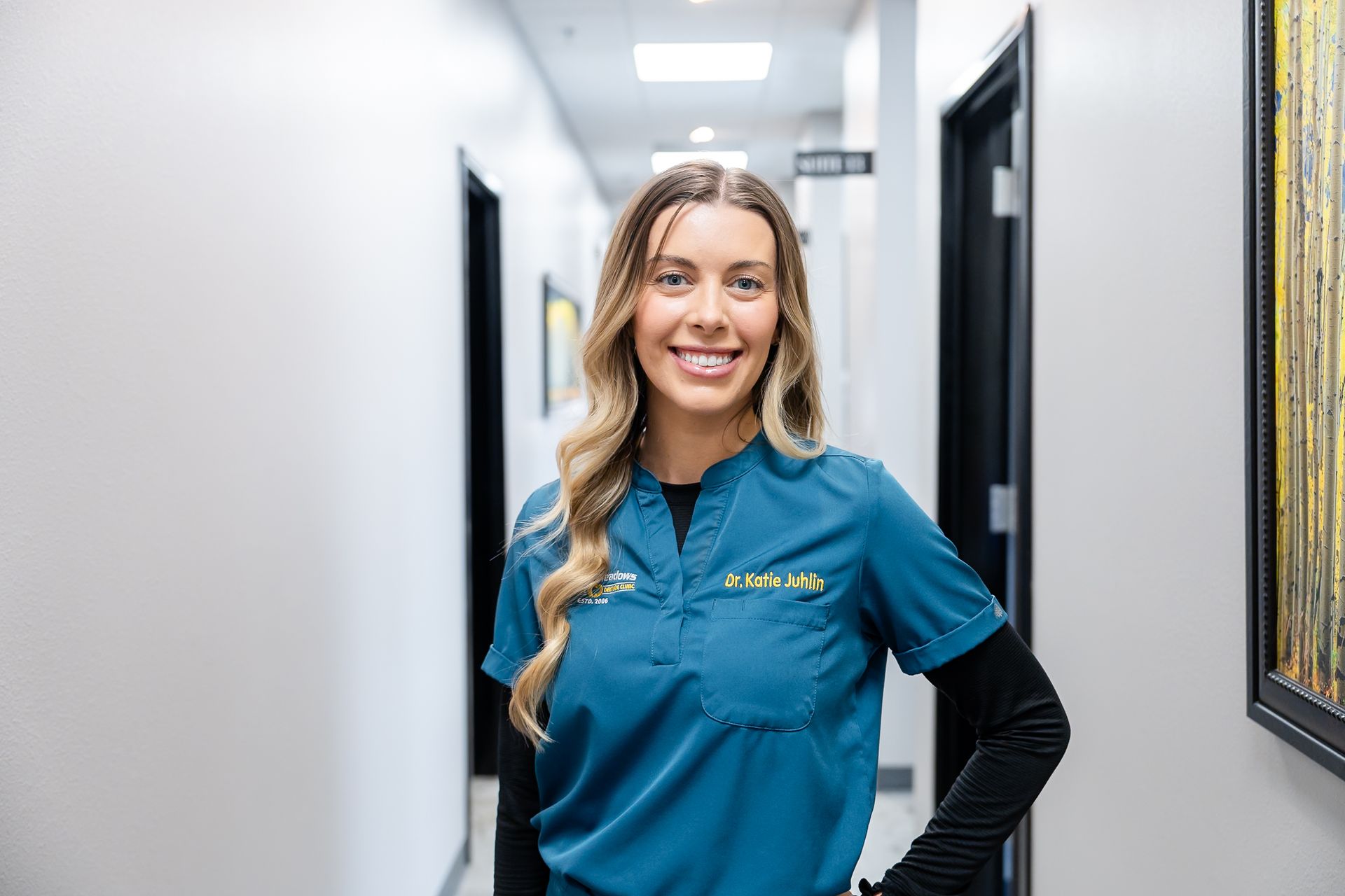 Dr. Katie in teal scrubs and a black long-sleeved shirt smiling in a bright office hallway.