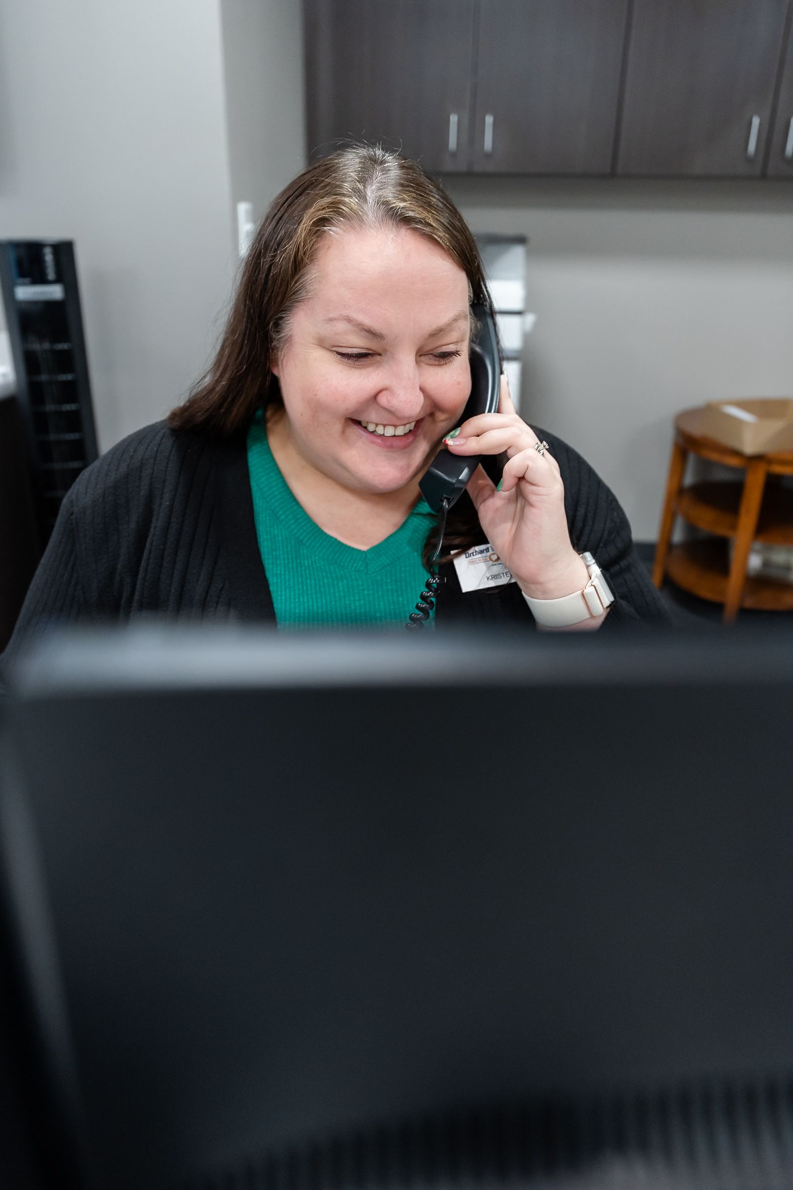 A smiling person wearing a green shirt and black cardigan sits at a desk talking on a telephone in an office.