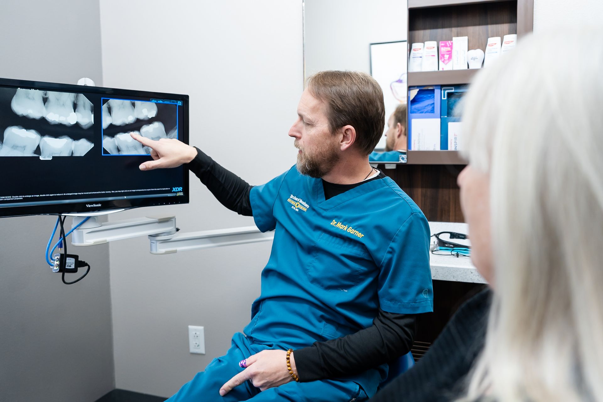 A dental professional speaks with a patient seated in a chair in a clinical office setting.