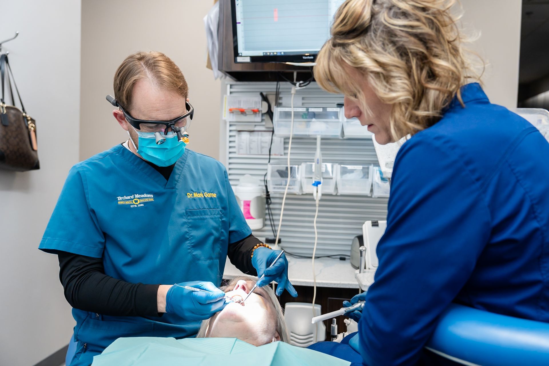 Dr. Garner and his assistant wearing scrubs and masks perform a procedure on a patient in a clinical dental office.