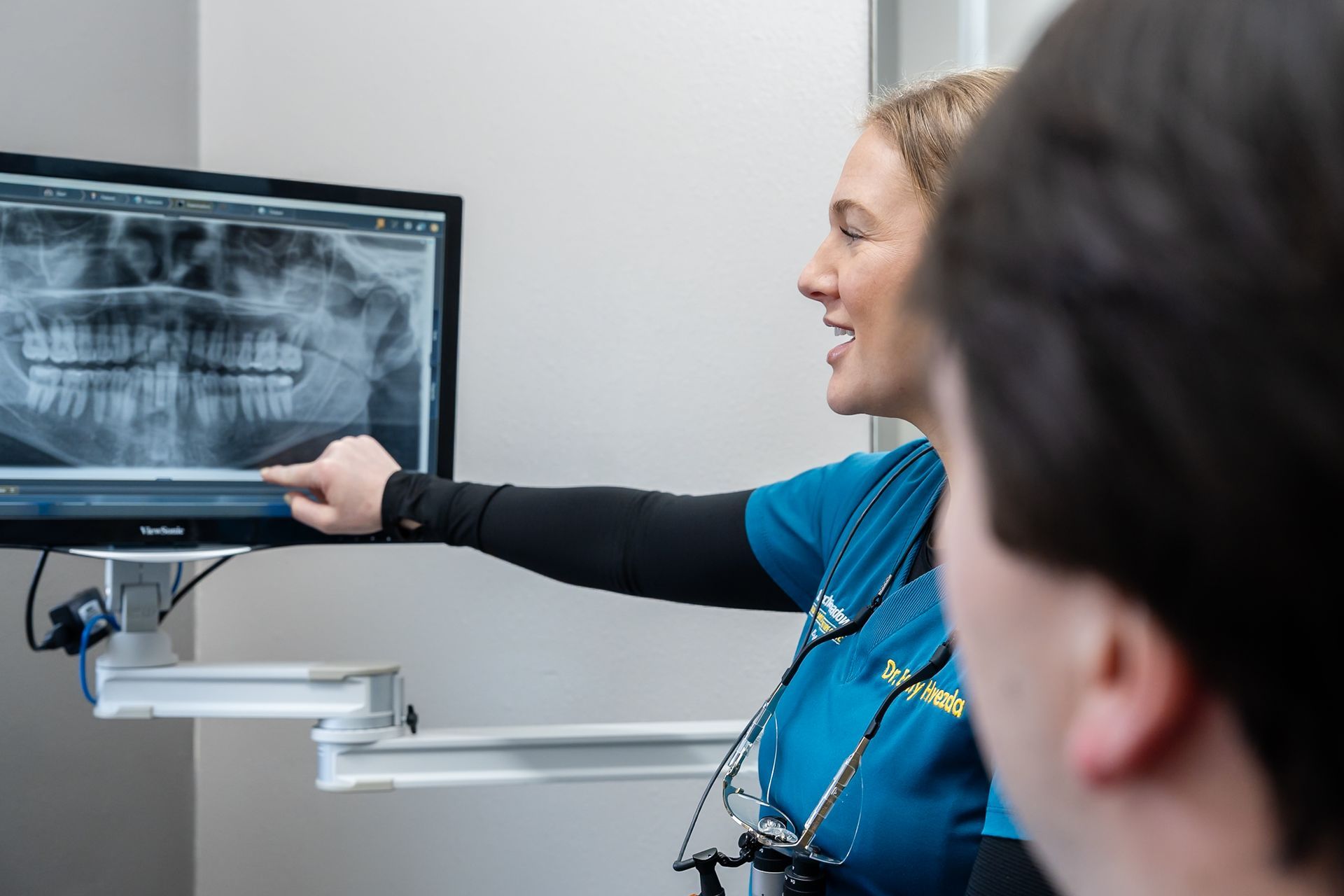 A dental hygienist in a teal scrub top discusses a tooth image on a monitor with a patient in a dental chair.