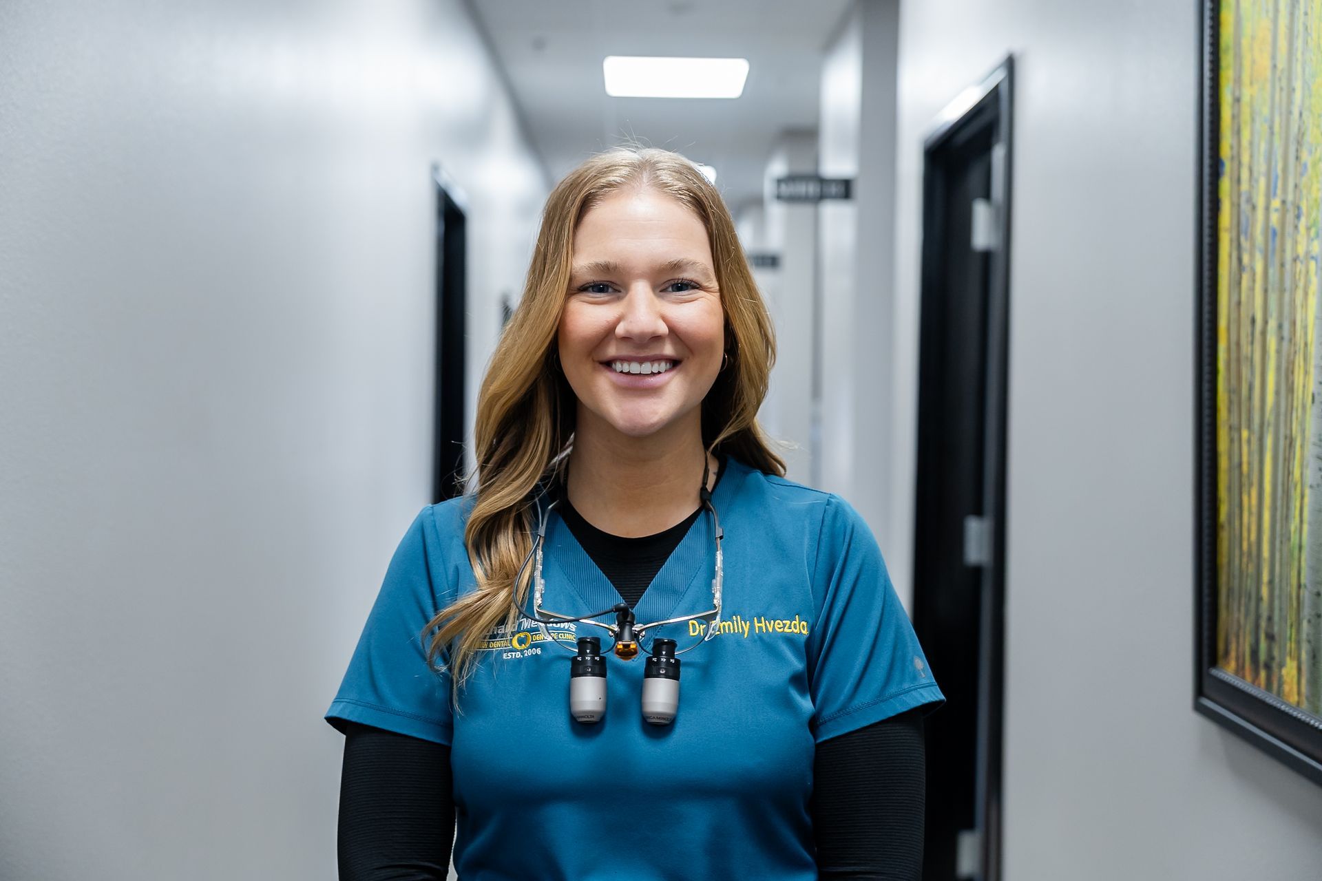 Dr. Emily in blue medical scrubs stands in a clinic hallway, wearing surgical loupes around their neck.