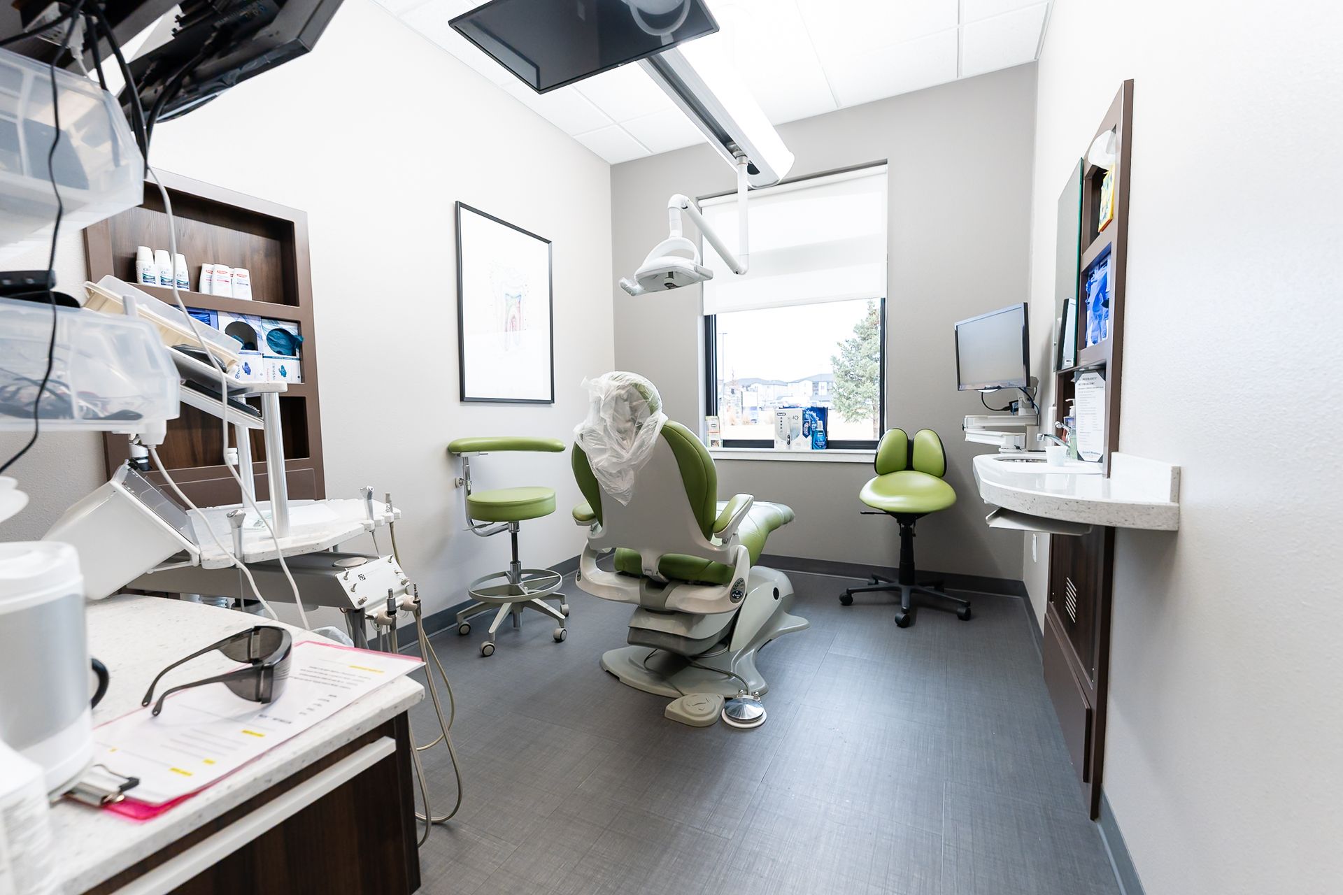 A modern dental exam room with a central patient chair, green swivel stools, and medical equipment in a clean, bright space.