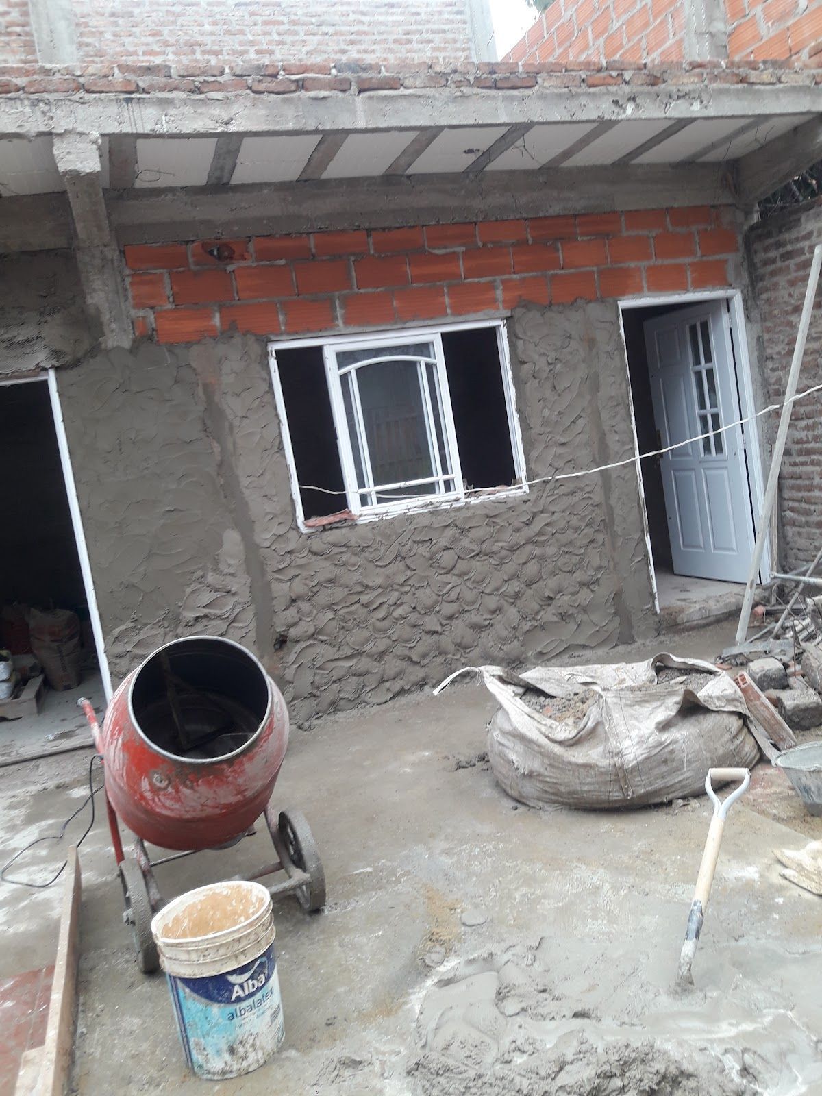 Herramientas de construcción y ladrillos apilados en un suelo polvoriento junto a una pared parcialmente enlucida.