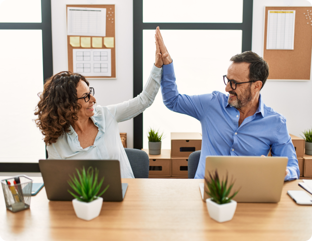Two coworkers high-five over laptops in a bright office setting, smiling at each other.