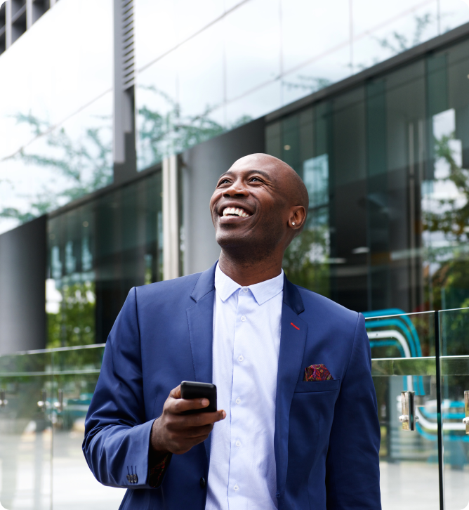 Smiling Black man in a blue suit holds a phone, looking upwards outside a modern building.