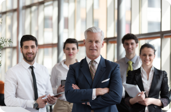 A group of five professionals in business attire, standing in an office setting; the central figure has crossed arms.