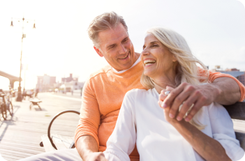 Smiling elderly couple sitting on a bench on a boardwalk. The man has his arm around the woman, they appear happy.