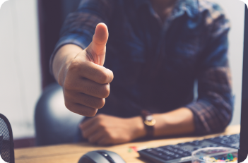Person giving a thumbs-up at a desk. They are wearing a blue plaid shirt and have a watch on their wrist.