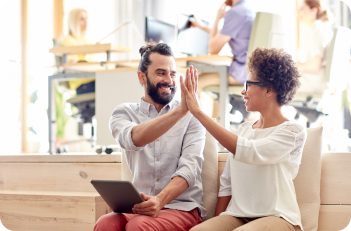 Two coworkers high-five. A man with a beard holds a tablet, smiling. A woman smiles, wearing glasses, in an office setting.