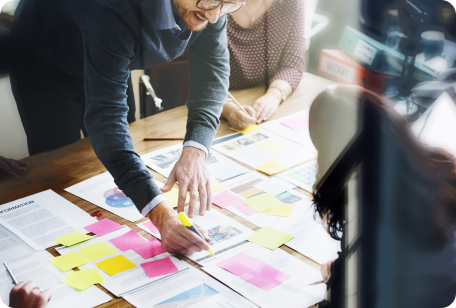 Man in glasses points at documents on a table with sticky notes, collaborating with colleagues.