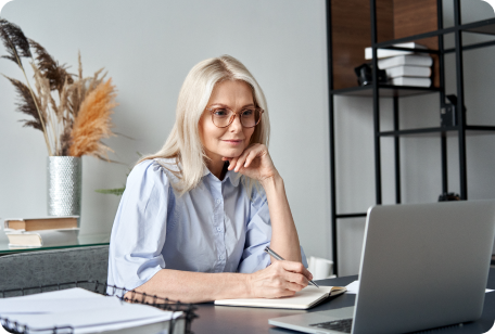 Blonde woman wearing glasses, working on a laptop at a desk. She is taking notes in a notebook.