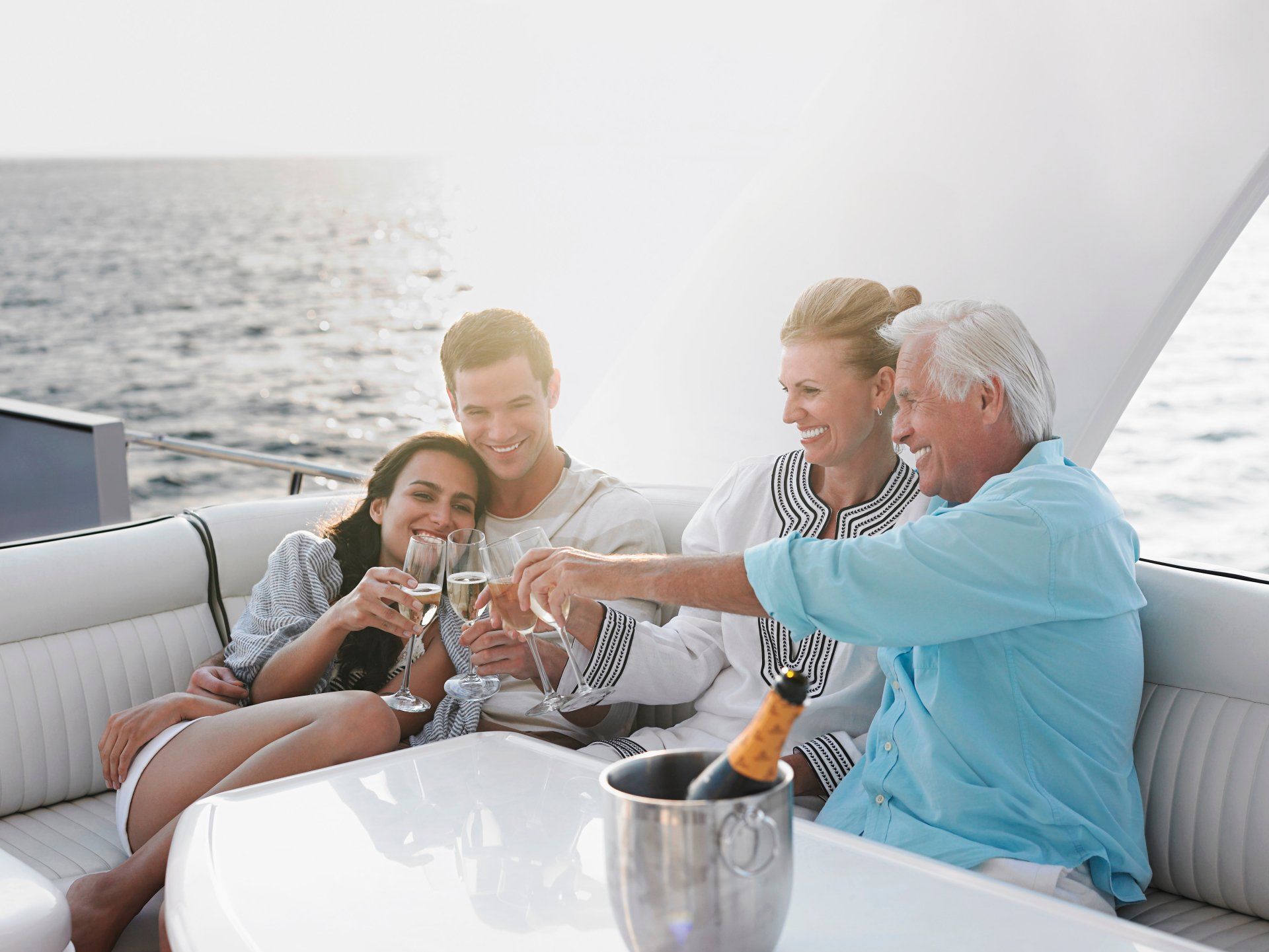 Four people on a yacht toasting with champagne. Smiling, they are outdoors with the ocean in the background, sunny setting.