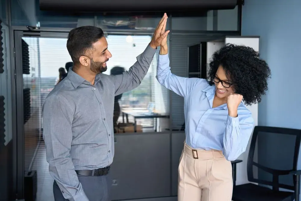 Two people high-fiving in office setting, smiling. One in blue shirt and khakis, the other in a gray shirt.