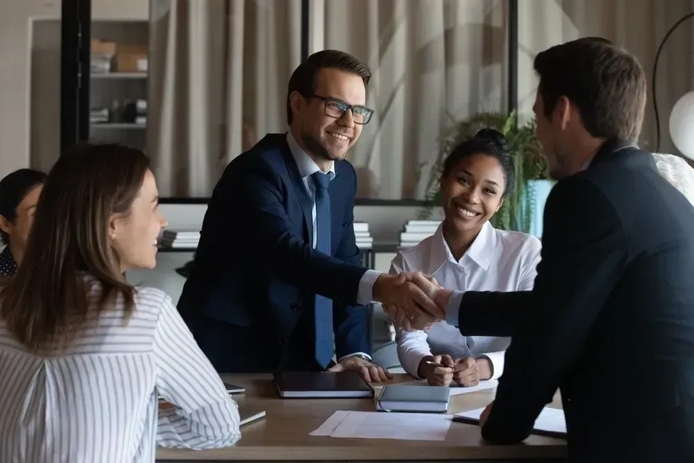 Three men in an office, one elderly, shaking hands with a younger man; other man holds papers.