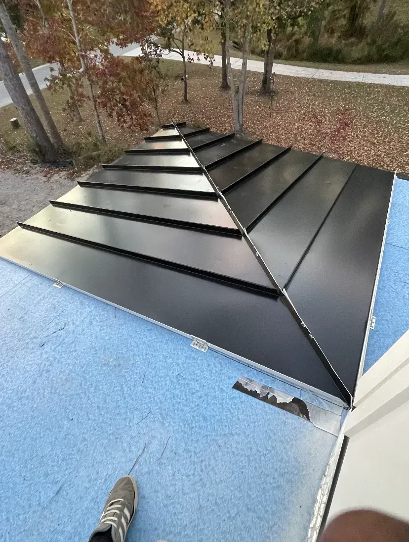 Black metal rooftop sections on a blue roof, viewed from above with trees in the background