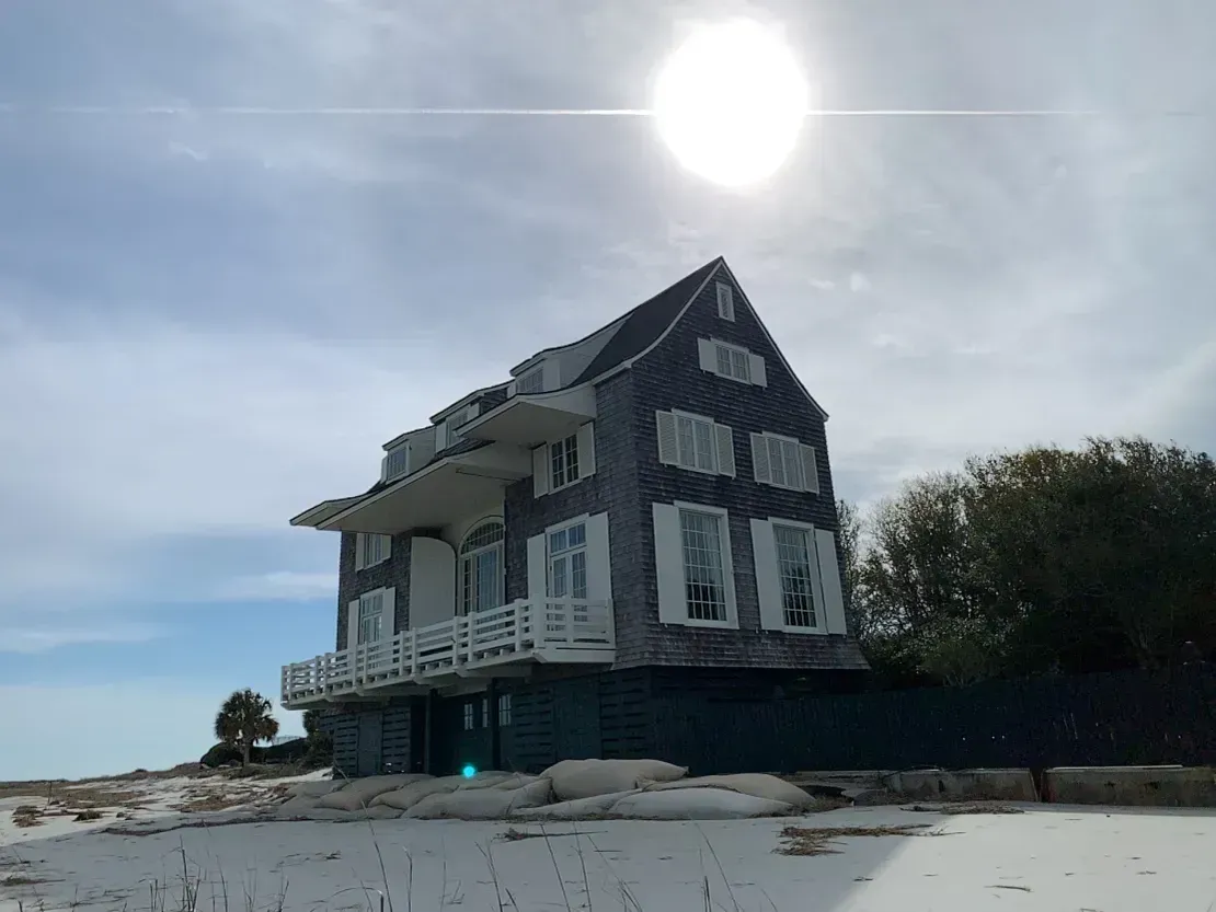 Gray beach house on a sandy shore under a bright sun, with calm water and a clear sky