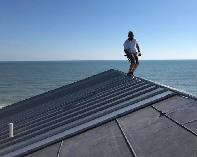 Person standing on a sloped metal roof overlooking the ocean under a clear blue sky