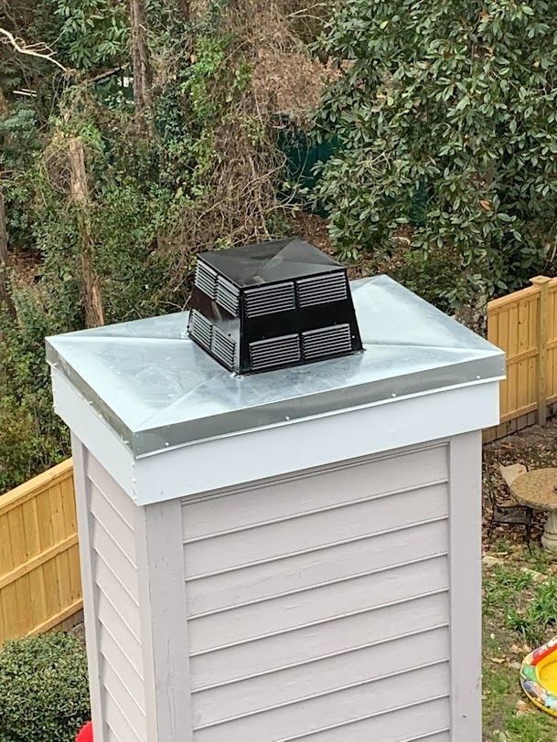 White vented utility box with a black chimney cap on a backyard fence line.