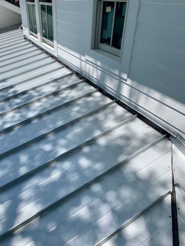 White metal rooftop deck beside house siding and windows, with sunlit shadows across the panels.