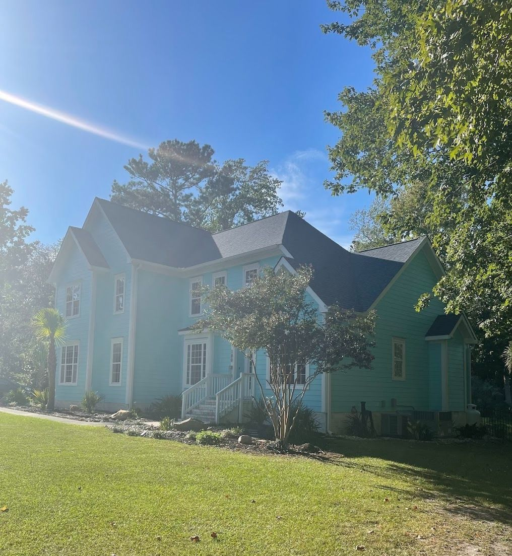 Light blue house with a dark roof on a sunny lawn, framed by trees and bright sky.