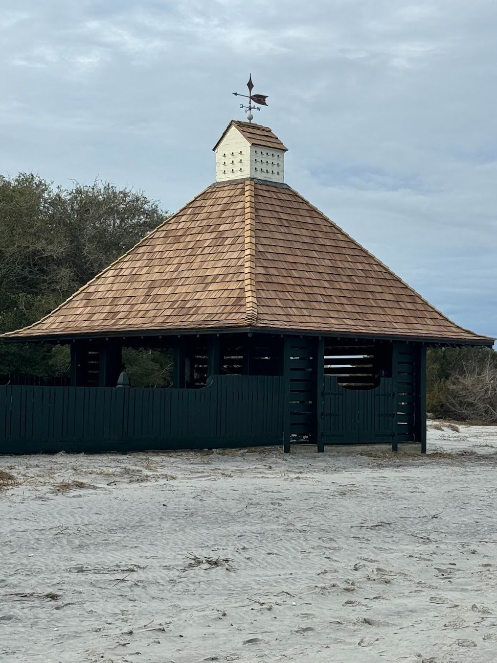 Small octagonal wooden pavilion with a shingled roof and weather vane, surrounded by gravel and trees