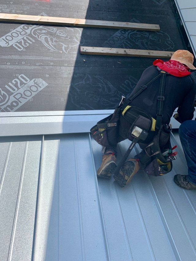 Worker in safety gear on a sloped metal roof beside a skylight, with roofing materials nearby.